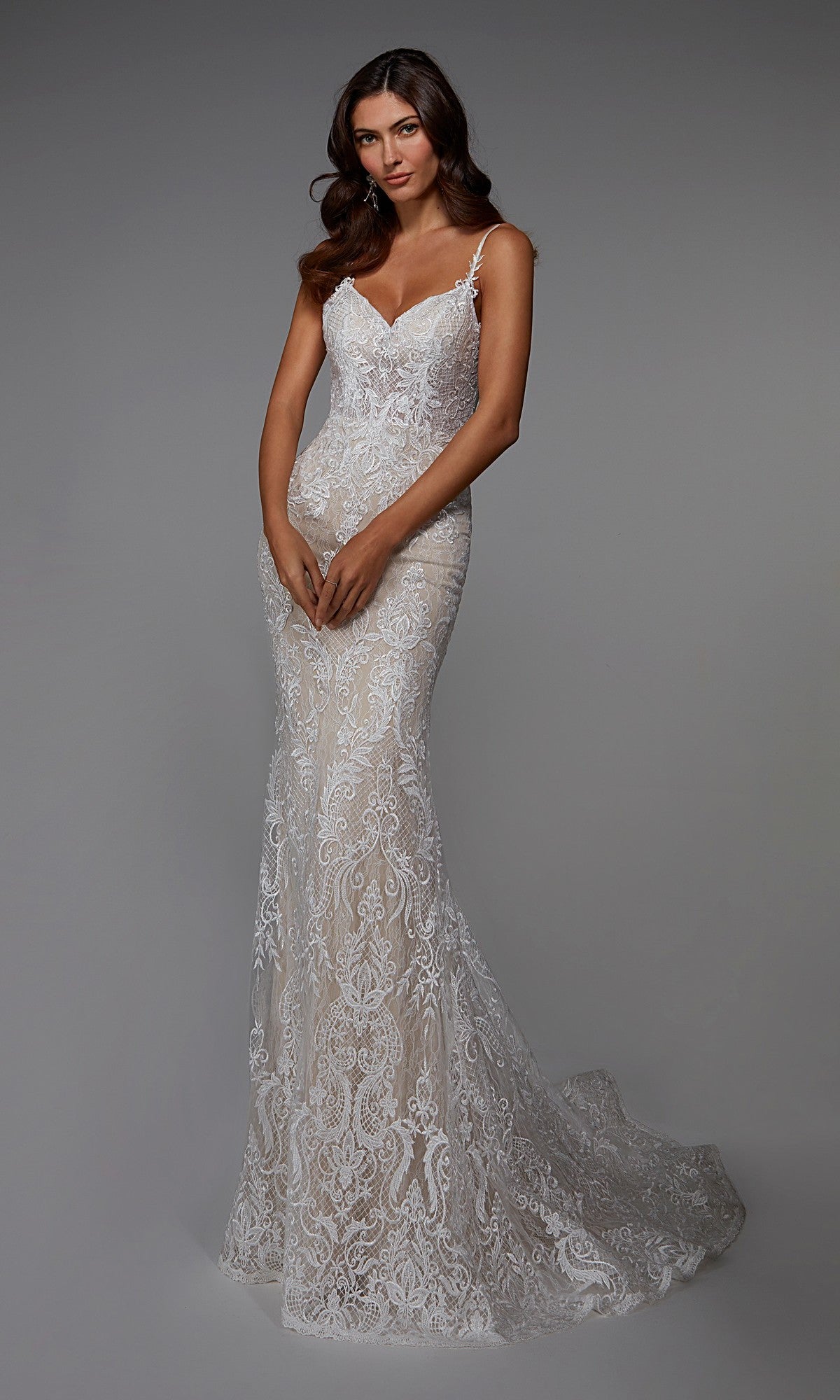 A woman models the Alyce 7028 long white dress in ivory/sand, a fitted, sleeveless gown with delicate lace embroidery and thin straps, standing against a plain gray background—an elegant choice for formal occasions.